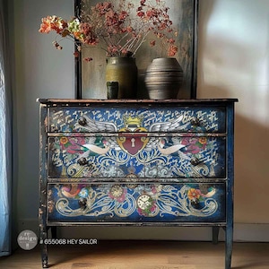 May include: A three-drawer blue chest of drawers with a distressed finish. The drawers are decorated with a winged heart, floral, and clock designs. The text "HEY SAILOR" is visible on the bottom left.