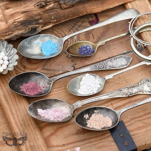 May include: Six antique silver spoons, each containing a different coloured powder: blue, purple, pink, white, light pink, and tan. The spoons are arranged on a wooden surface, with a decorative metal dish and bracelets visible.