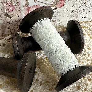 May include: Three wooden spools with white lace wrapped around them. The spools are brown and have a rough texture. The lace is delicate and has a floral pattern.