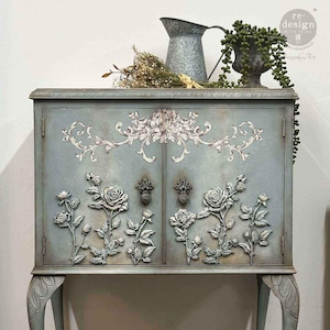 May include: A vintage-style cabinet in a muted blue-green hue, adorned with ornate floral relief carvings and decorative hardware. The cabinet features a detailed floral design across the doors and sides, with a distressed finish. A metal pitcher and a potted plant sit on top.