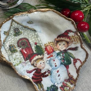 May include: A decorative, shell-shaped dish with a gold-painted rim. The dish features a winter scene with two elves building a snowman in front of a stone house. The elves wear red and green outfits and hats. The background is white with snow.