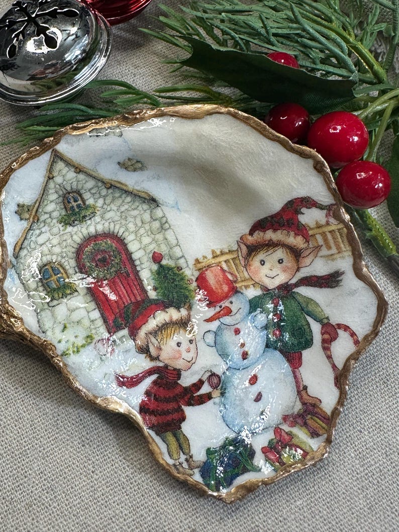 May include: A decorative, shell-shaped dish with a gold-painted rim. The dish features a winter scene with two elves building a snowman in front of a stone house. The elves wear red and green outfits and hats. The background is white with snow.