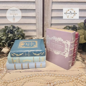 May include: A stack of three antique-style books in shades of blue, with gold accents and ornate designs. A larger, lavender-colored book stands beside them. The books are arranged on a white surface with a string of pearls.
