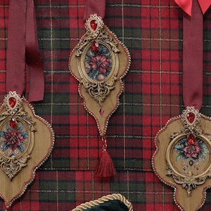 May include: Ornate, wooden Christmas ornaments with red jewel accents and floral centerpieces hang against a red, green, and black plaid background. Each ornament has a burgundy ribbon for hanging and a decorative tassel.