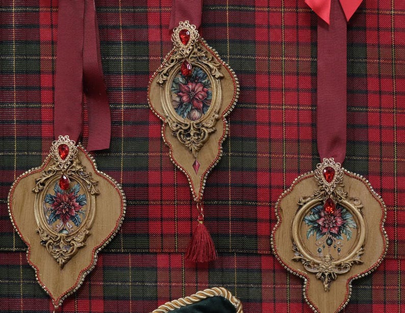 May include: Ornate, wooden Christmas ornaments with red jewel accents and floral centerpieces hang against a red, green, and black plaid background. Each ornament has a burgundy ribbon for hanging and a decorative tassel.