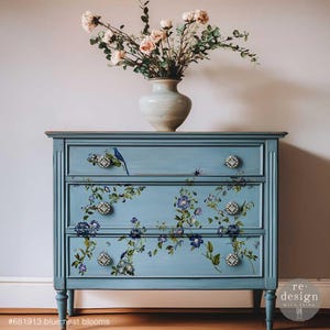 May include: A light blue painted dresser with three drawers, adorned with floral decals and bird accents. The dresser has decorative knobs and is topped with a vase of pink flowers. The text "#681913 blue-nest blooms" is visible at the bottom.