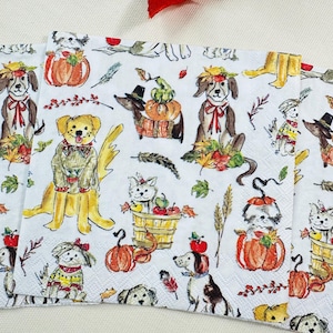 May include: White paper napkins with a repeating pattern of dogs dressed in fall attire. The dogs are wearing hats, scarves, and sweaters, and are surrounded by pumpkins, leaves, and apples. The napkins are perfect for a fall-themed party.