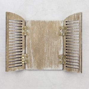 Puede incluir: Decoración de pared de madera envejecida que simula una ventana con contraventanas. Las contraventanas están abiertas, revelando un panel central. La pieza está pintada en un color neutro con vetas de madera visibles y bisagras metálicas.