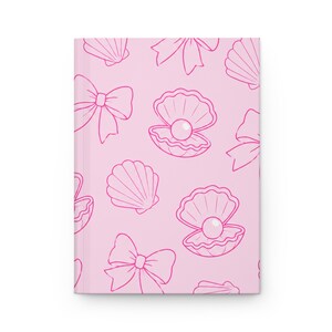 May include: A light pink hardcover journal featuring a repeating pattern of dark pink line art illustrations. The design includes open seashells containing pearls, closed seashells, and decorative bows distributed across the front cover.