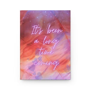 May include: A hardcover journal with a colorful abstract design in shades of pink, orange, and purple. The cover features the text "It's been a long time coming" in a handwritten style, with small heart shapes scattered across the design.