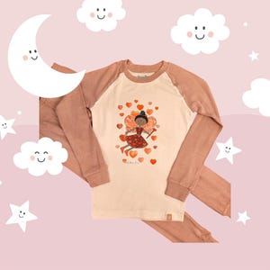 May include: A two-piece pajama set in beige and mauve. The long-sleeved top features a graphic of a fairy with heart accents. The pajamas are displayed on a pink background with cartoon clouds, stars, and a crescent moon.