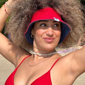 May include: A red bucket hat with a navy blue brim is worn by a person. The person is also wearing a red bikini top and a gold necklace. The person is smiling and looking to the side. The background is a beach.
