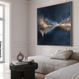 Op de afbeelding: Groot abstract schilderij met een gouden lijst. Het kunstwerk heeft een donkerblauwe achtergrond met gouden en blauwe accenten, waardoor een reflecterend, landschapachtig ontwerp ontstaat. Het schilderij hangt boven een lichtgekleurde bank in een moderne woonkamer.