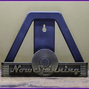 May include: A gold-colored metal record holder with the words "Now Spinning" on the front. The holder is designed to hold a vinyl record and has a black plastic stand behind it.