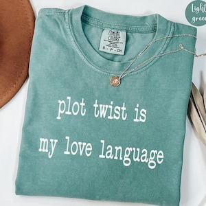 May include: Light green t-shirt with the text "plot twist is my love language" in white. A gold necklace with a pendant is draped over the shirt. The shirt is a Comfort Colors brand.