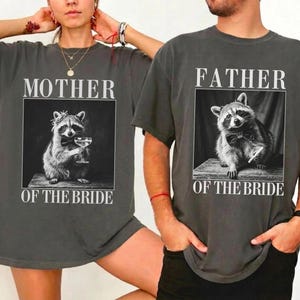 May include: Two charcoal gray t-shirts. One shirt reads "MOTHER OF THE BRIDE" above a black and white image of a raccoon holding a glass. The other shirt reads "FATHER OF THE BRIDE" above a similar raccoon image.