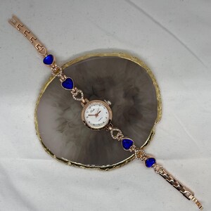 May include: A gold-tone wristwatch with a white face and blue heart-shaped accents on the band. The watch is on a white surface.