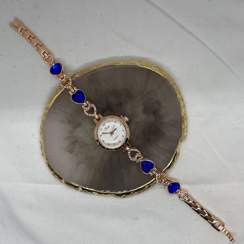 May include: A gold-tone wristwatch with a white face and blue heart-shaped accents on the band. The watch is on a white surface.