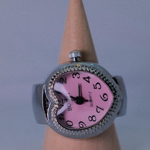 May include: A silver heart-shaped ring watch with a pink face and black hands. The watch face has black numbers and the words "QUARTZ" and "BOLTZ" printed on it.