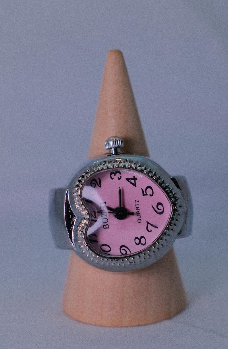 May include: A silver heart-shaped ring watch with a pink face and black hands. The watch face has black numbers and the words "QUARTZ" and "BOLTZ" printed on it.