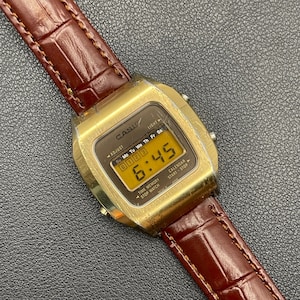 May include: A gold-toned Casio digital watch with a brown leather band. The watch face displays the time as 6:45. The band has a textured, crocodile-like pattern. The watch has the text "CASIO" on the face.