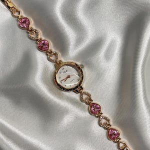 May include: A gold-tone wristwatch with a white face and black Roman numeral hour markers. The watch has a pink crystal heart-shaped band.