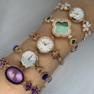 May include: Five gold-tone wristwatches with different face designs and crystal accents. The watches have a variety of colored face designs, including green, white, and purple.
