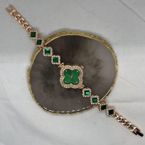 May include: A gold-tone watch with a green face and a clover-shaped design. The watch has a gold-tone band with square green gemstones.