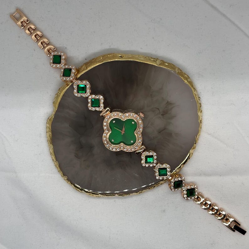 May include: A gold-tone watch with a green face and a clover-shaped design. The watch has a gold-tone band with square green gemstones.