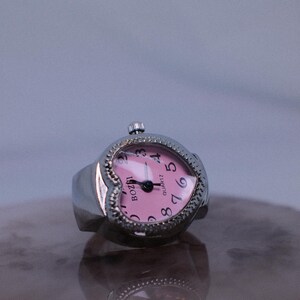 May include: A silver heart-shaped ring watch with a pink face and black hands. The watch face has black numbers and the text "QUARTZ" and "BOZZINI".