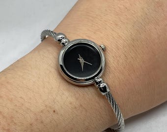 May include: A silver bangle watch with a black face and silver hands. The watch has a twisted band.