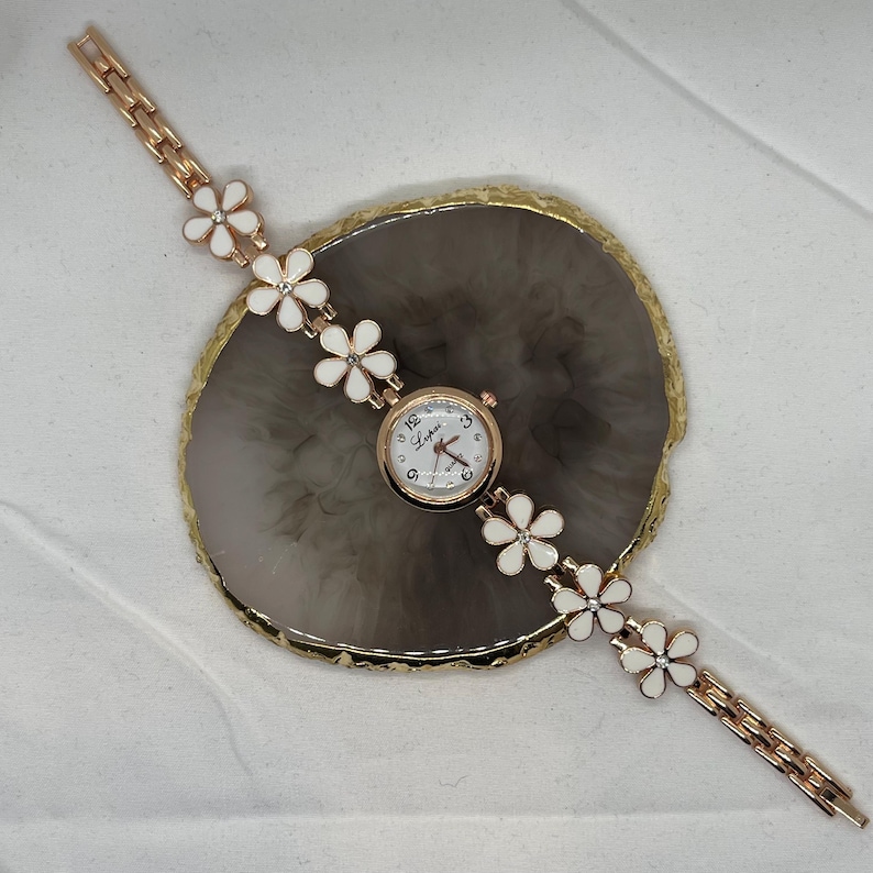 May include: A gold-tone wristwatch with a white face and white flower accents on the band.