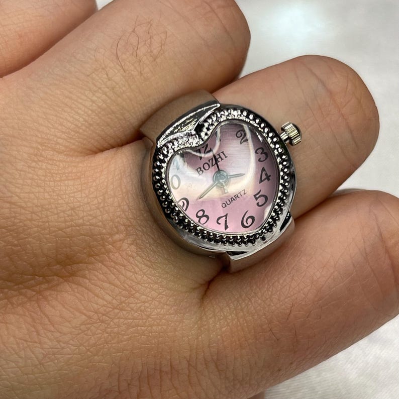 May include: A silver heart-shaped ring watch with a pink face. The watch face has black numbers and the word "BOZHI" printed on it. The watch is set to 10:10.