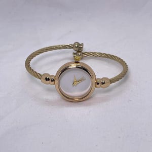 May include: A gold-toned bangle watch with a white face and gold hands. The watch band is made of a braided metal.