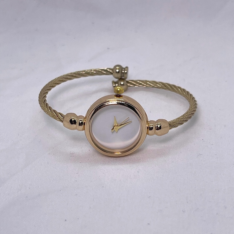 May include: A gold-toned bangle watch with a white face and gold hands. The watch band is made of a braided metal.
