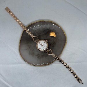 May include: A rose gold wristwatch with a white face and roman numeral hour markers. The watch has a heart-shaped design on the band and is on a light gray background.