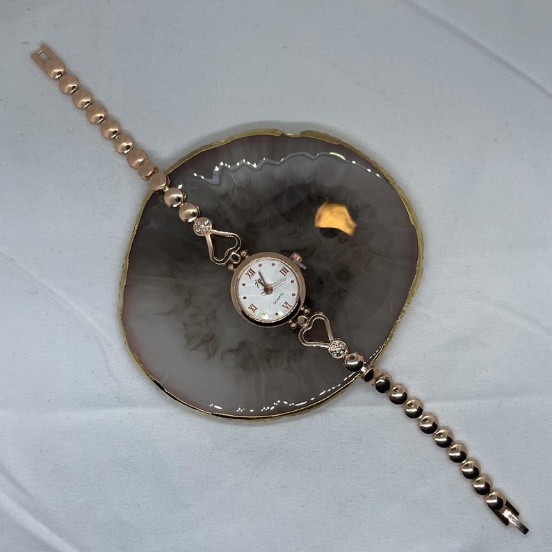 May include: A rose gold wristwatch with a white face and roman numeral hour markers. The watch has a heart-shaped design on the band and is on a light gray background.