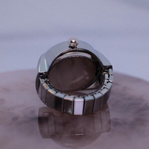 May include: A silver watch with a white band. The watch is closed and the back of the watch is visible.