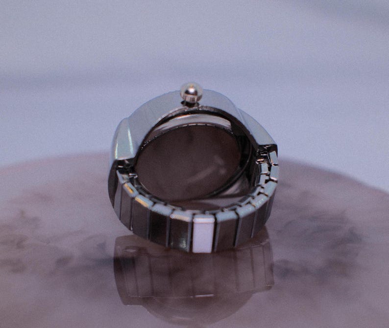 May include: A silver watch with a white band. The watch is closed and the back of the watch is visible.