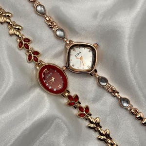 Puede incluir: Dos relojes de pulsera dorados con correas decorativas. Uno tiene una esfera ovalada roja con números romanos, el otro tiene una esfera cuadrada blanca. Ambos relojes tienen la marca "Zido" en la esfera y están sobre un fondo blanco.