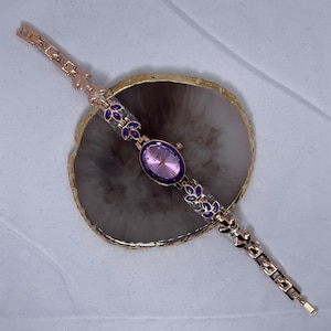 May include: A gold-tone wristwatch with a pink face and purple accents on the band. The watch is on a gray surface.
