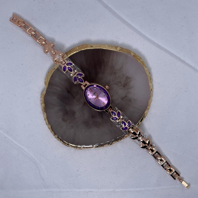 May include: A gold-tone wristwatch with a pink face and purple accents on the band. The watch is on a gray surface.