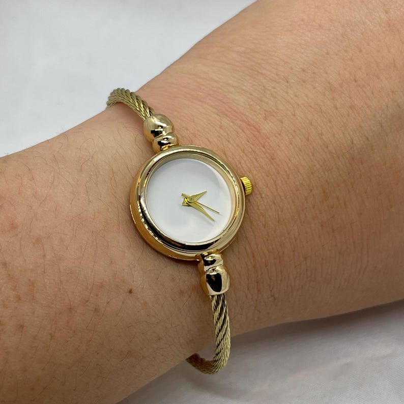 May include: A gold-toned bangle watch with a white face and gold hands. The watch has a cable-style band.