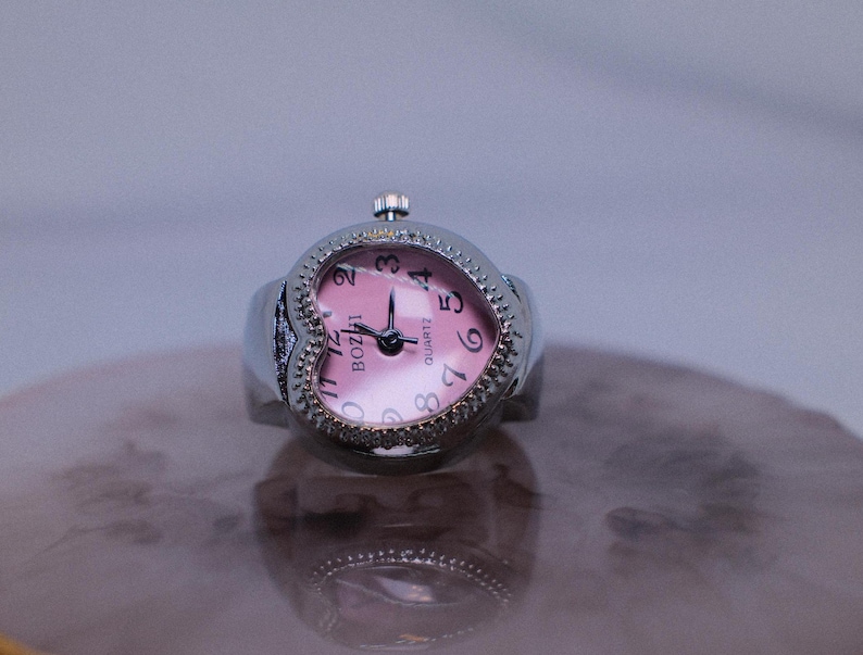 May include: A silver heart-shaped watch with a pink face and black numbers. The watch is set to 12:30 and says "BOZII" QUARTZ on the face.