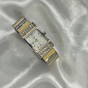 Puede incluir: Un reloj de cuarzo rectangular con una correa plateada y una esfera blanca. El reloj presenta una correa color crema con detalles de pedrería y una esfera blanca con números negros. Es un accesorio elegante.