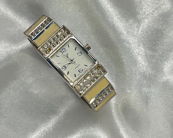 May include: A rectangular quartz watch with a silver band and a white face. The watch features a cream-colored band with rhinestone accents and a white watch face with black numerals. The watch is a stylish accessory.