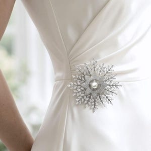 May include: A sleeveless ivory dress featuring a V-neckline and a gathered waist detail. Attached to the waist is a large, circular silver-toned brooch shaped like a starburst, adorned with numerous clear crystals and a single central white pearl.