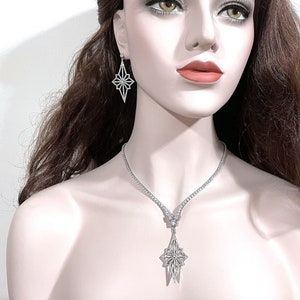 May include: A silver-toned necklace and earring set featuring a starburst or geometric diamond-shaped pendant and matching drop earrings. The necklace consists of a delicate chain with a central star-shaped charm, all encrusted with clear stones.