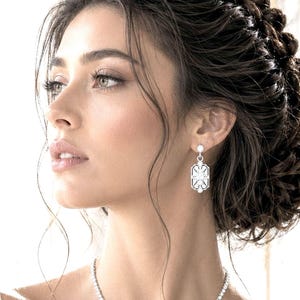 May include: A silver-toned drop earring featuring an elongated octagonal frame with geometric cutouts and a central clear stone. The earring is suspended from a small circular stud, showcasing a symmetrical design with clean lines and metallic finish.