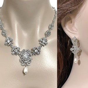 May include: A silver necklace with a large, ornate pendant featuring a pearl drop. Matching earrings with a pearl drop dangle from silver filigree.